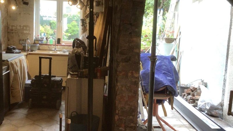 Kitchen stripped back to bare plaster and boards during the works, with tools and materials stacked against the walls